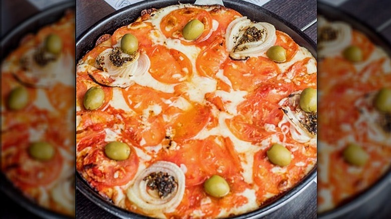 A full pizza covered in cheese, tomatoes, onions, lives, and seasoning sits in a pan.