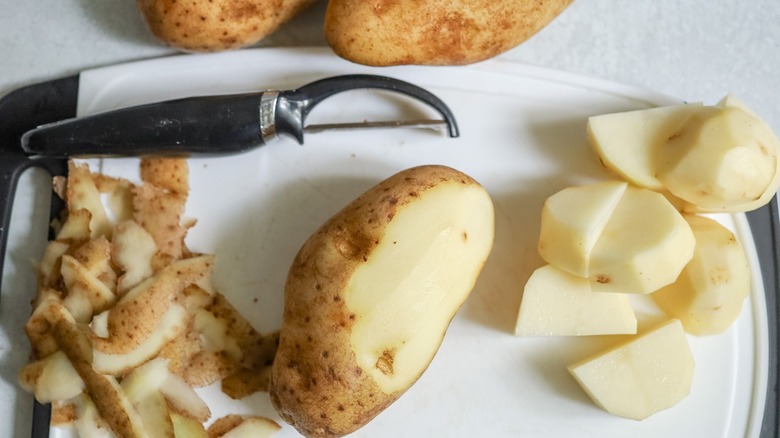 Semi-peeled russet potato on cutting board with peeler and sliced potato chunks