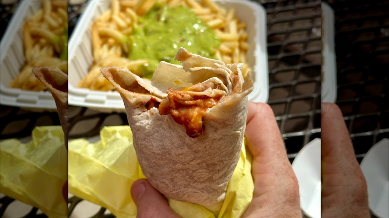 A hand holding a bean and cheese burrito with red sauce in front of fries with guacamole