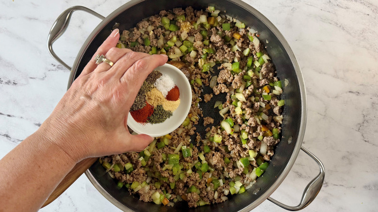 Hand holding small bowl of spices over ground meat and diced vegetable mixture in skillet