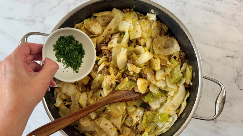 Hand holding small bowl of minced parsley over skillet with dirty cabbage