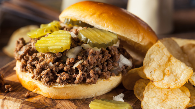 A Maid-Rite on cutting board next to potato chips. The bun is off, revealing pickles and onions on top.