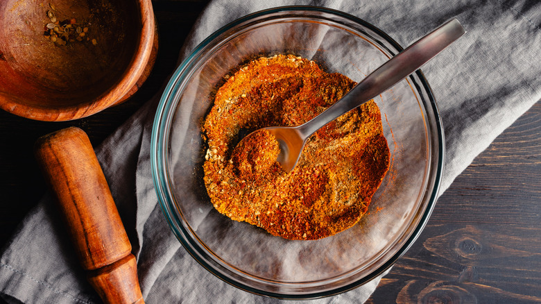 Spoon in glass bowl of mixed spicy seasonings next to mortar and pestle on gray cloth.