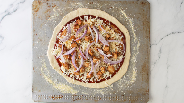 unbaked pizza crust with red sauce, chicken, onions, and cheese on a sheet pan