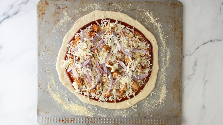 unbaked pizza crust with red sauce, chicken, onions, and cheese on a sheet pan