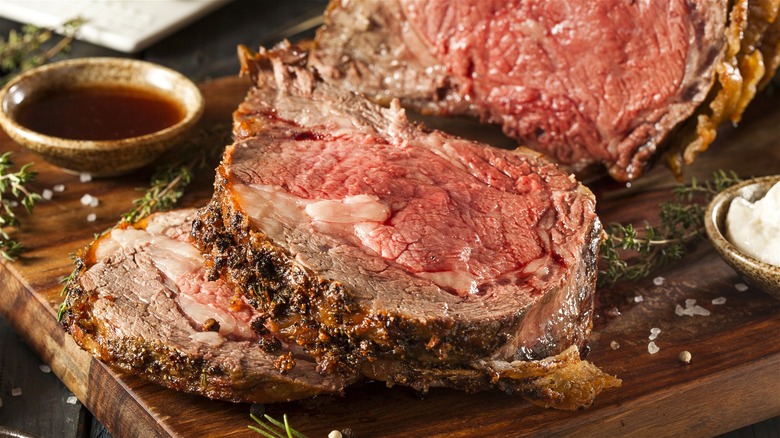 Slices of prime rib on a wooden board, with sauce and seasonings on the side.