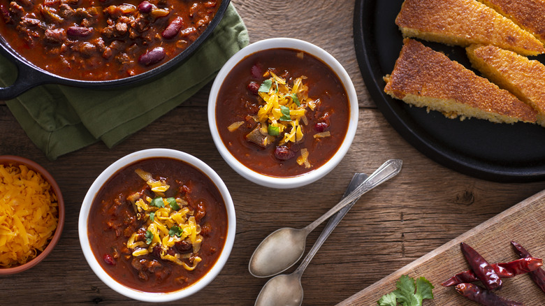 Two bowls of chili with toppings on a table with cornbread and the remaining pot of chili