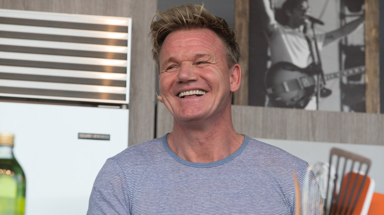 Gordon Ramsay in a light blue t-shirt smiling in front of a gray wall with a pictures of a musician