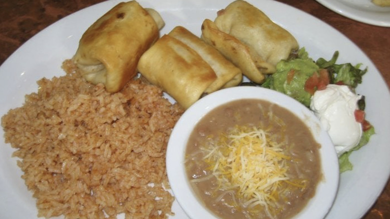 A plate with three chimichangas, Mexican rice, refried beans, sour cream, and guacamole