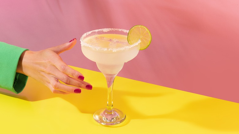 Person reaching for classic margarita with salted rim and lime garnish on yellow table with pink background