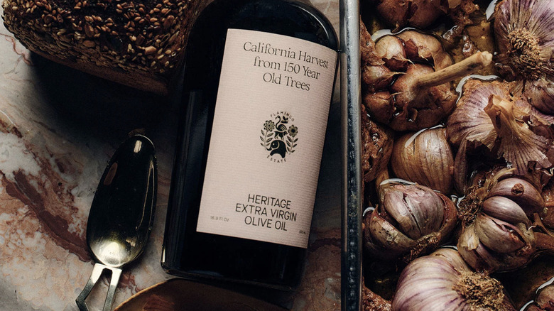 Bottle of Heritage Extra Virgin Olive Oil with loaf of seeded bread and tray of garlic cloves.