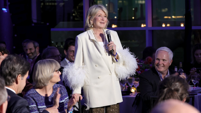 Martha Stewart wearing a white feathered blazer while speaking into a microphone at an event