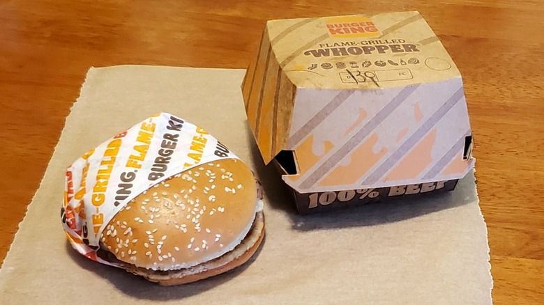 a Burger King Whopper in wrapper next to its takeout box on table