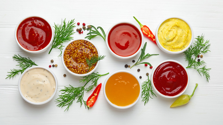 A variety of dipping sauces in ramekins arranged with peppers and herbs on a white board.