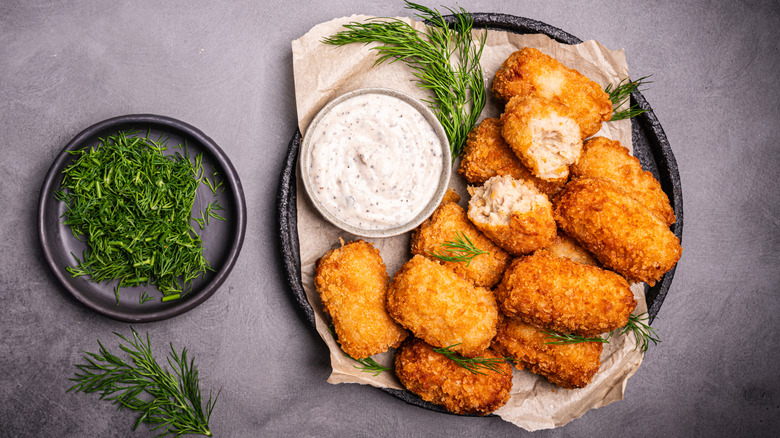 Croquettes with sauce and fresh herbs on a brown napkin on a black plate with a side plate of fresh dill