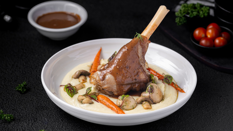 Lamb shank in white bowl over mashed potatoes, carrots, and mushrooms.