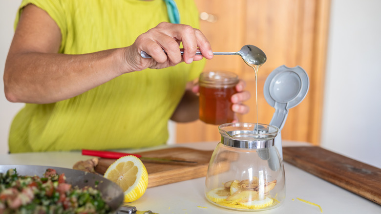 Person holding a glass honey jar and scooping honey with a metal spoon into a tea kettle