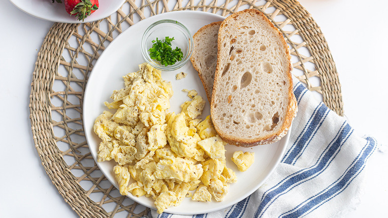 Overhead shot of a plate of scrambled eggs next to a couple of slices of sourdough bread and a small glass cup of chives