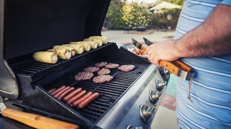 Man standing beside grill with hot dogs, burgers, and corn