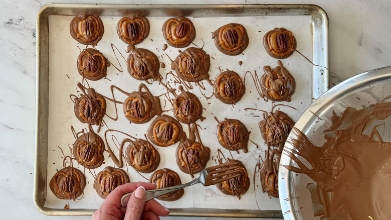 Hand using fork to drizzle chocolate over turtles on baking sheet