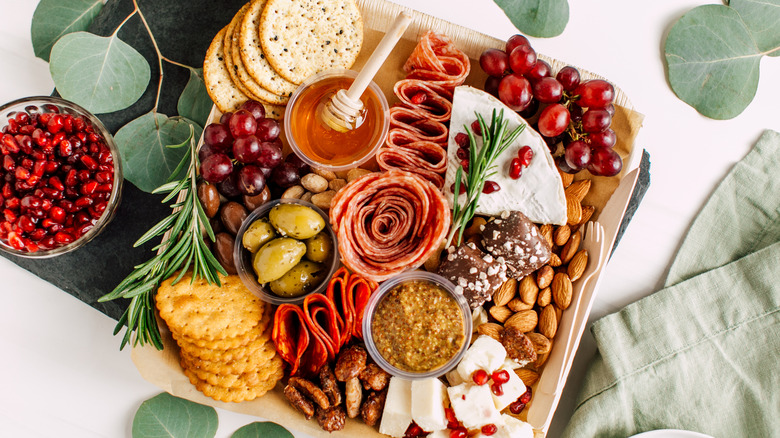 charcuterie board with crackers, honey, olives, pomegranate seeds, grapes, nuts, mustard, meats, and cheeses on white background with pale green leaves