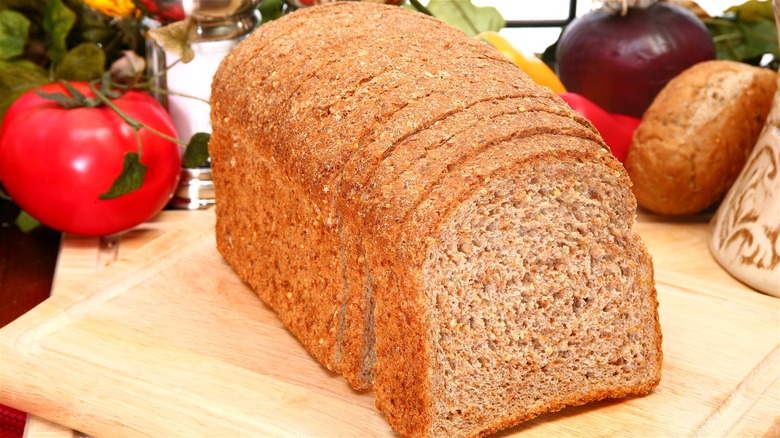 A sliced loaf of grain bread on a wooden cutting board, with assorted produce in the background.