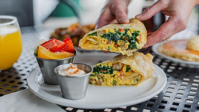 Hand holding half of a breakfast burrito with a side of fruit