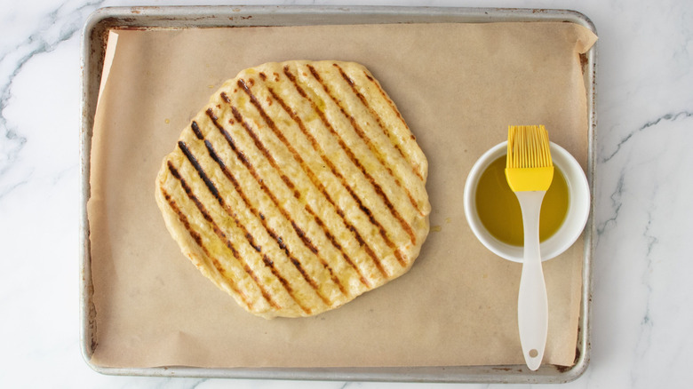Grilled pizza crust and bowl of oil with pastry brush on baking sheet