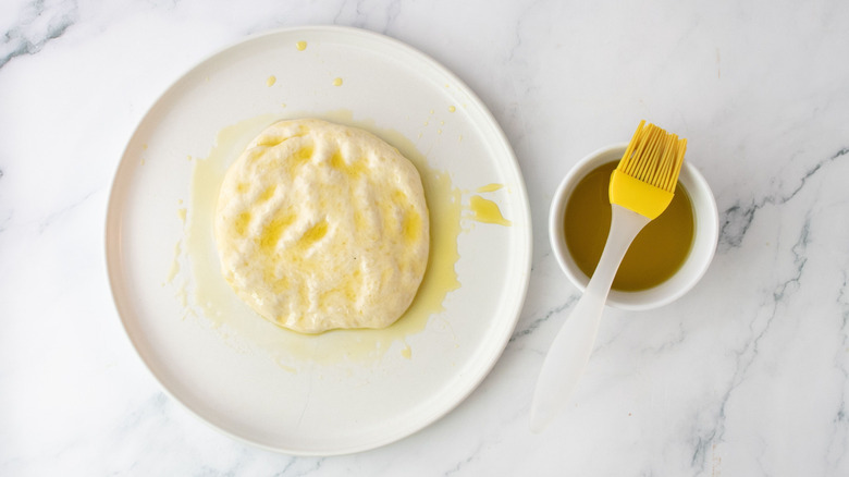 Oil-coated pizza dough on plate next to bowl with pastry brush