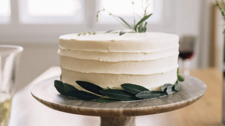 A cake covered in white frosting and decorated with leaves sits on a cake stand