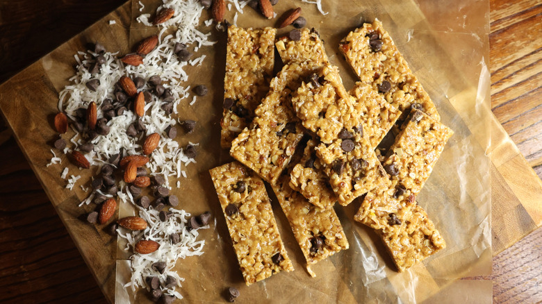 overhead view of a pile of no-bake cereal bars on wax paper on a cutting board next to dried coconut, chocolate chips, and almonds