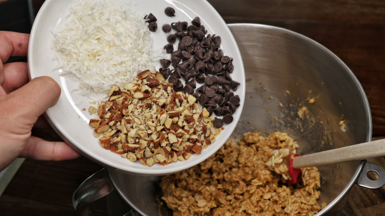 hand tilting a plate of chopped almonds, dried coconut, and chocolate chips above a mixing bowl of cereal bar mixture