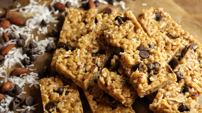 pile of cereal bars on wax paper surrounded by dried coconut, chocolate chips, and almonds