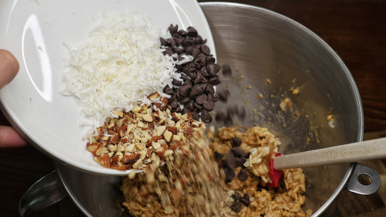 chopped almonds, chocolate chips, and dried coconut flakes pouring off a plate into a mixing bowl full of cereal bar mixture