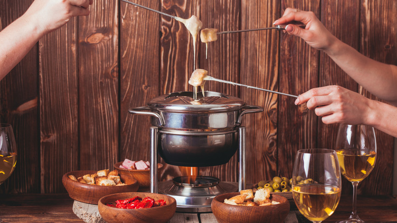 People dipping bread cubes into a fondue pot with bowls of bread, olives, peppers, and glasses of white wine surrounding the pot