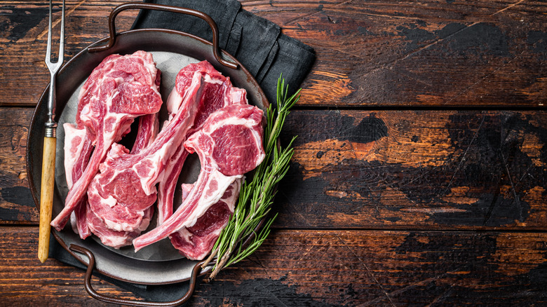 A pan filled with several raw lamb chops next to fresh herbs and a carving fork