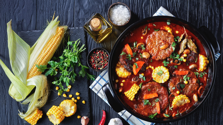 Burger patties basting in a tomato sauce with corn, carrots, and other veggies next to herbs and corn on the cob