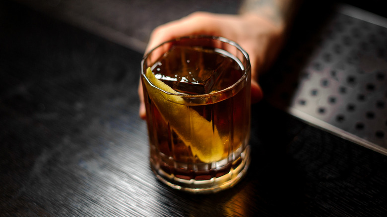 Hand holds rocks glass with brown liquor, ice cube, lemon twist