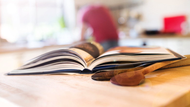 An open cookbook on a wooden kitchen surface with wooden spoons beside it