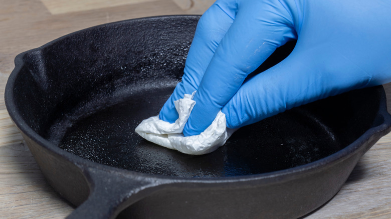 A hand seasoning a cast iron skillet with a paper towel and blue glove