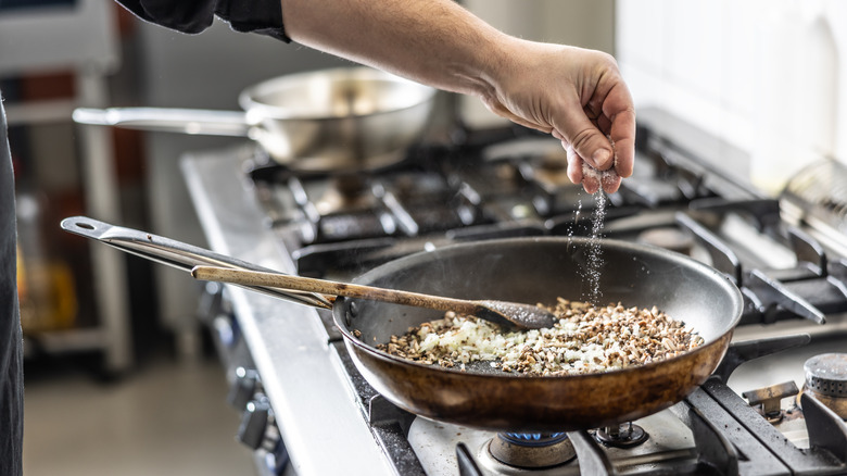 A man sprinkling salt into a frying pan filled with onions and mushrooms