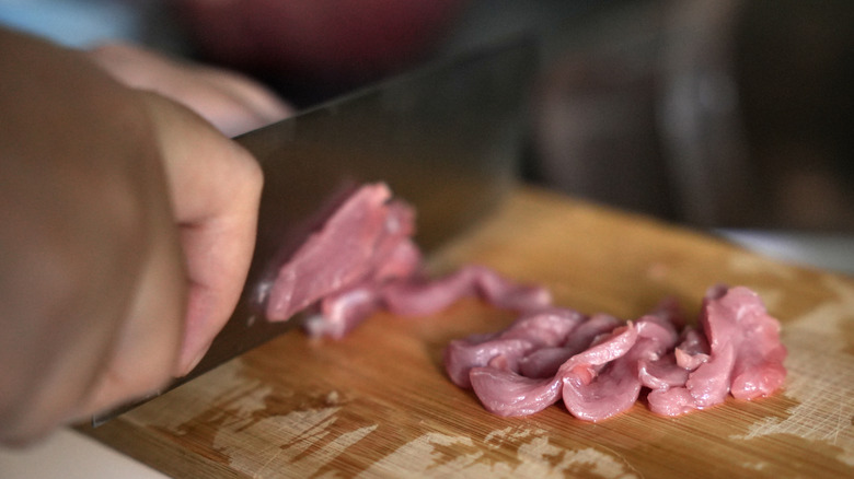 Hands cutting raw meat thinly with a knife on a wooden cutting board