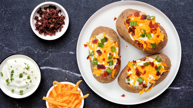 A plate of loaded baked potatoes next to bowls of chopped bacon, sour cream, and shredded cheese