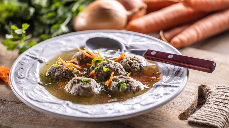 A bowl of dumplings in broth, served with a spoon, on a wood table with vegetables in the background