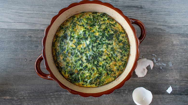 A dish filled with spinach soufflé next to a cracked eggshell