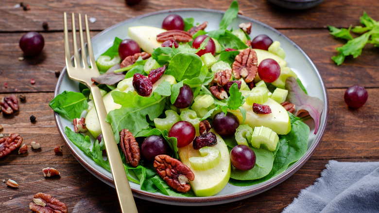 A plate of Waldorf salad with a fork on top resting on a wooden table that's sprinkled with grapes and walnuts