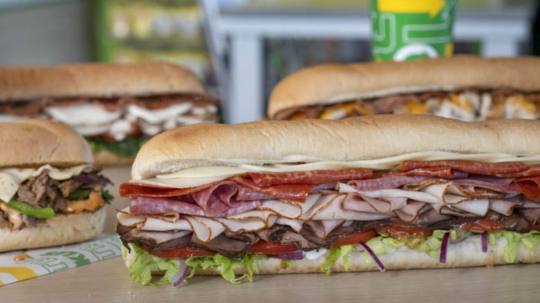 Four Subway foot longs on a table with a beverage cup in the background.