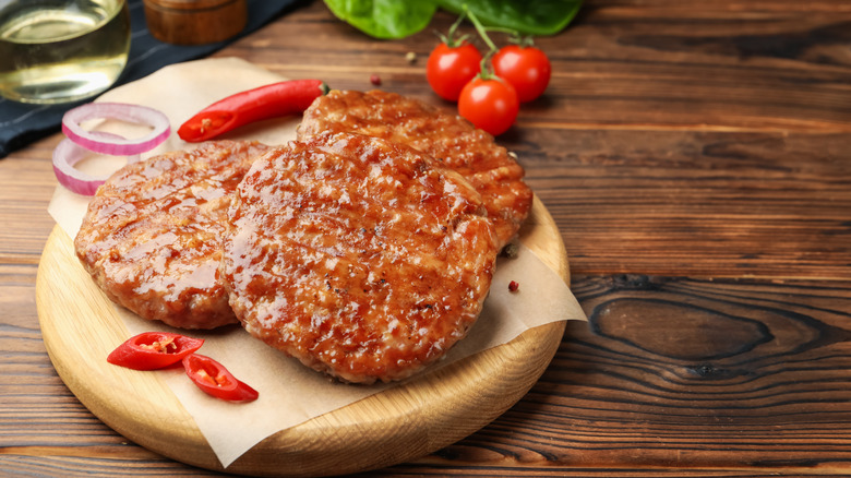 Three grilled hamburger patties on a wooden dish next to chopped chili, red onion, and whole tomatoes