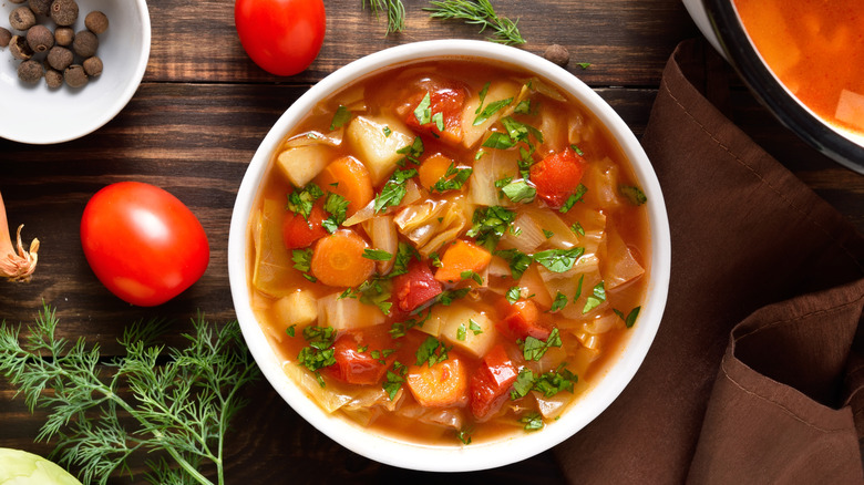 A bowl of vegetable cabbage soup next to whole tomatoes and herbs on a wooden tbale