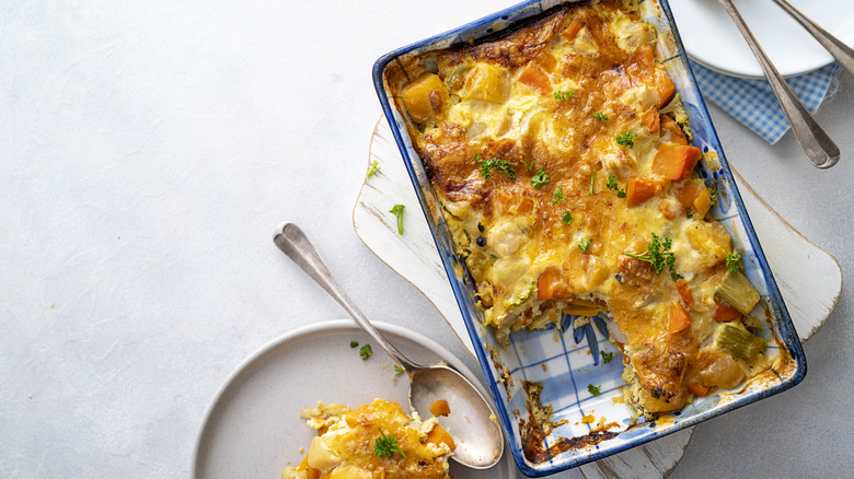 A casserole of carrots and other veggies in a baking dish, a serving scooped out onto a plate